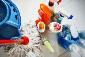 Closeup top view of unrecognizable home cleaning products with blue bucket and a mop on the side. All products placed on white tiled bathroom floor.