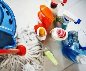 Closeup top view of unrecognizable home cleaning products with blue bucket and a mop on the side. All products placed on white tiled bathroom floor.